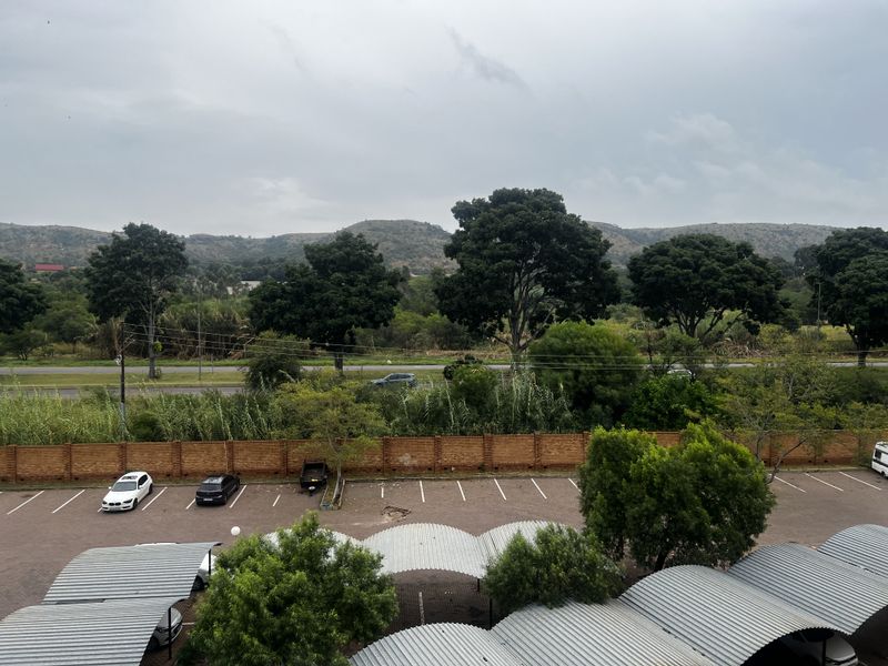 Elevated view overlooking a paved parking area with covered carports, a brick boundary wall, and a scenic landscape with trees and hills in the background.