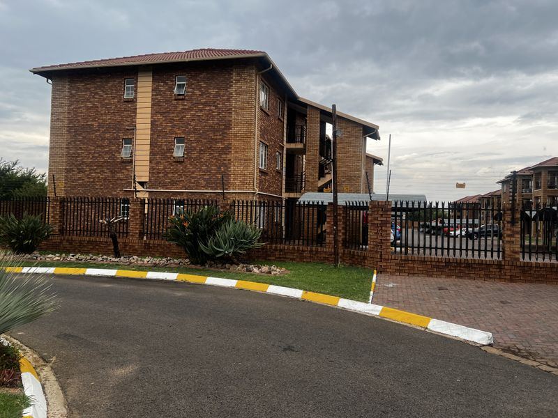 Exterior view of a multi-story brick apartment building featuring perimeter security fencing, paved driveway, and landscaped garden areas.