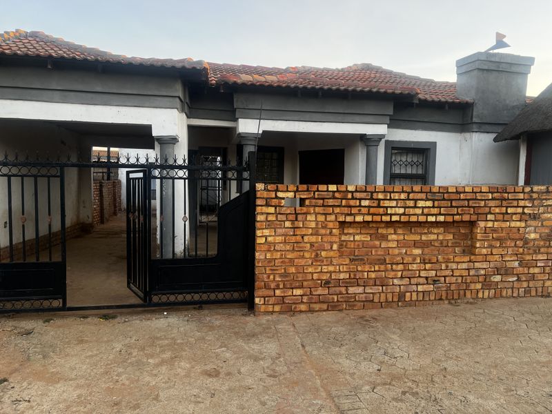 Front exterior view of a single-story house featuring a brick boundary wall, metal security gate, and covered entrance area with tiled roof.