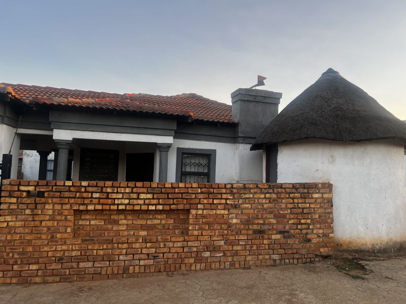 Front view of a house featuring a tiled roof section, a traditional thatched rondavel, and a face-brick boundary wall.