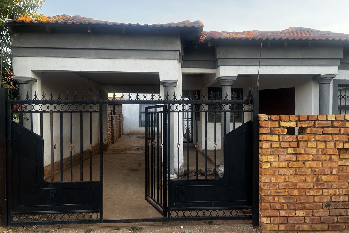 Front view of a single-story house featuring a covered entrance, decorative pillars, and a black metal security gate with a brick boundary wall.