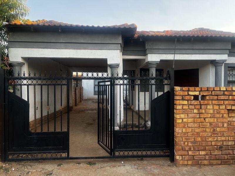 Front view of a single-story house featuring a covered entrance, decorative pillars, and a black metal security gate with a brick boundary wall.