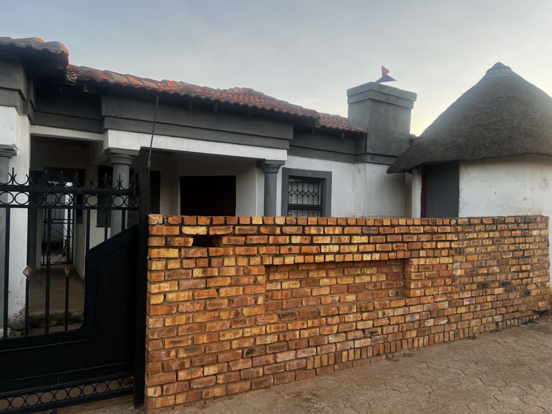 Front view of a residential property featuring a brick boundary wall, metal gate, covered entrance porch, and a traditional thatched roof structure.