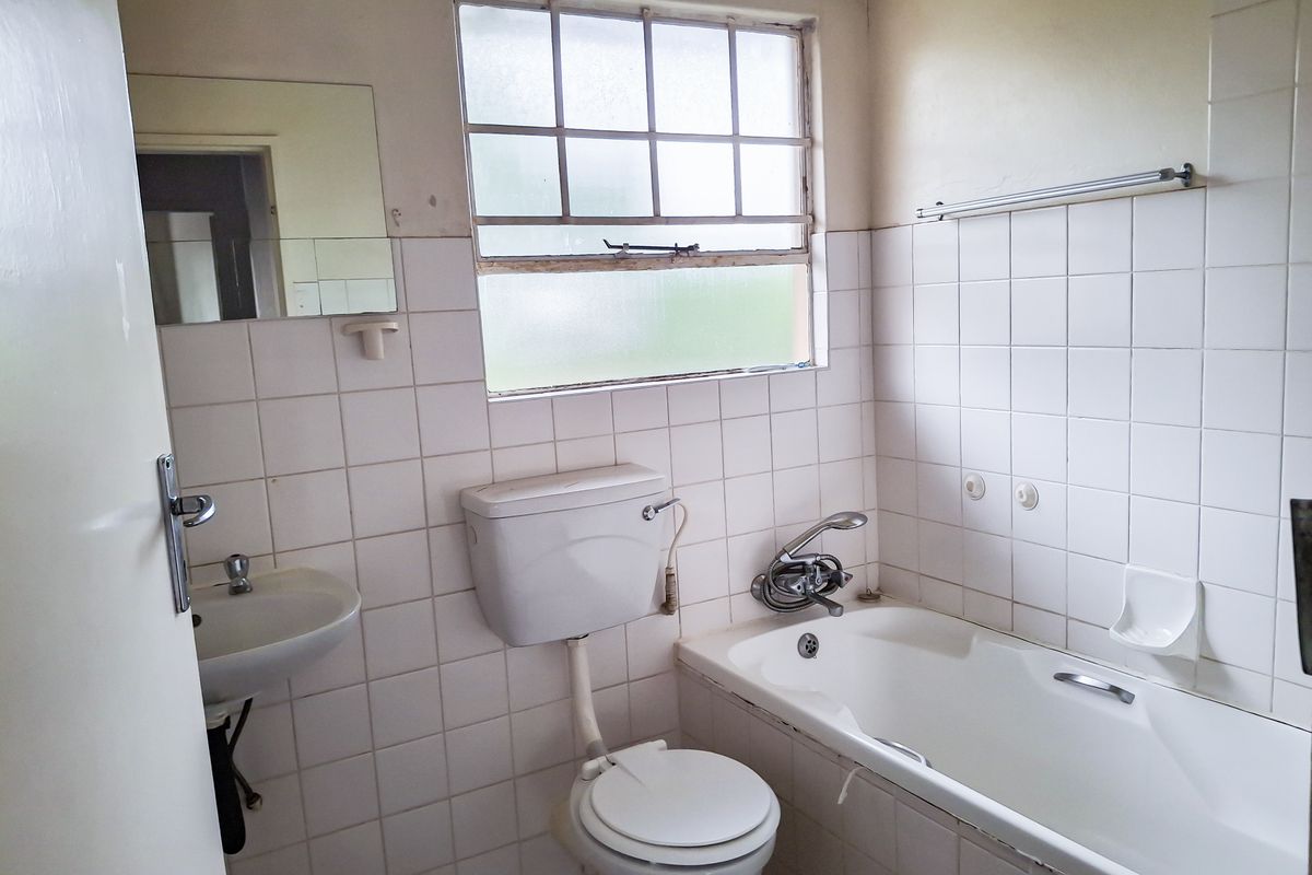 A dated bathroom featuring a standard bathtub, wall-mounted basin, and toilet with original white wall tiling and a metal-framed window.
