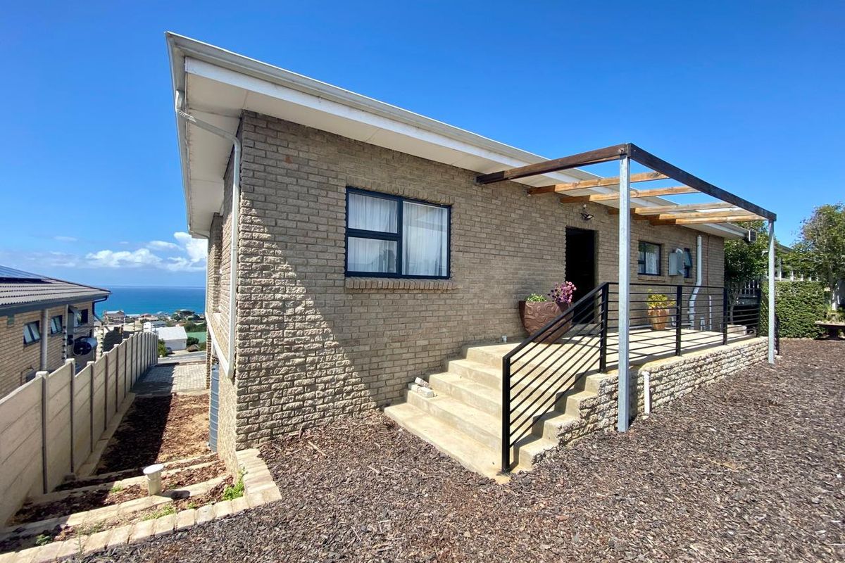 Exterior view of a brick house featuring a raised concrete patio with a timber pergola, metal railings, and ocean views in the distance.