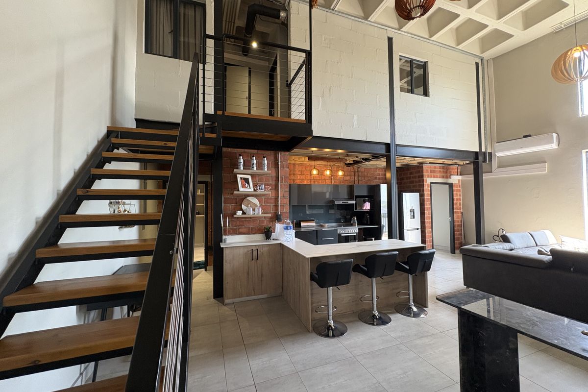 Modern open-plan apartment featuring a double-volume living area, industrial-style staircase, and a contemporary kitchen with a breakfast bar and exposed brick accents.