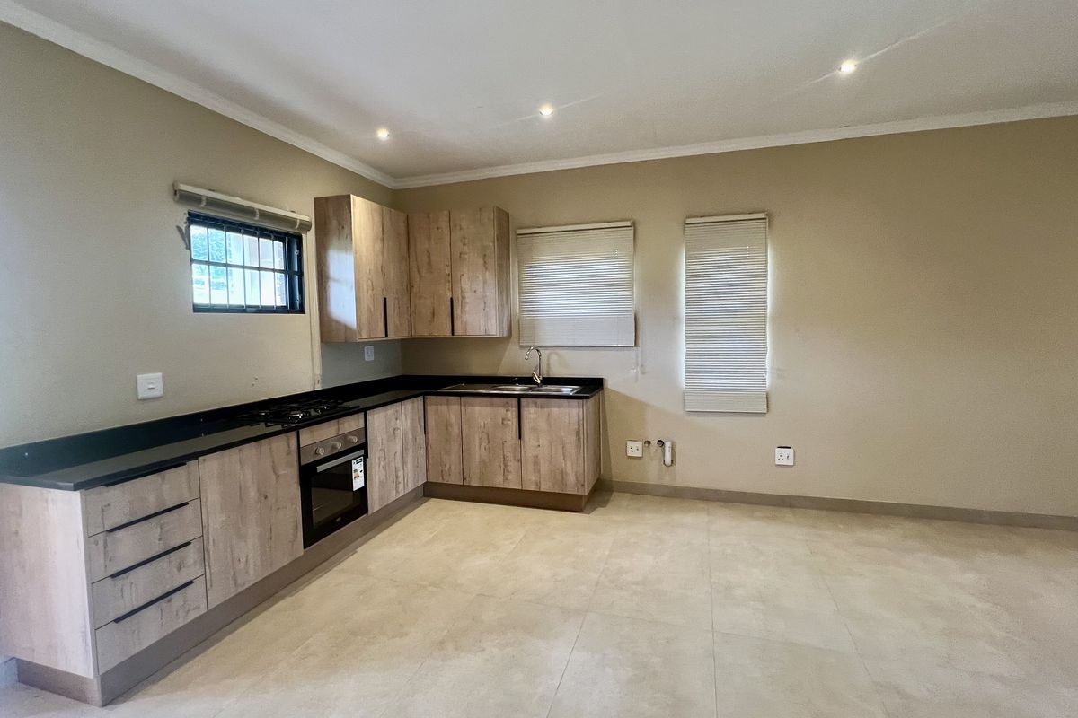 A clean, modern kitchen featuring light wood-grain cabinetry, dark countertops, built-in oven and hob, and neutral tiled flooring in an open-plan layout.