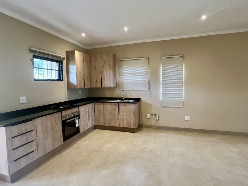 A clean, modern kitchen featuring light wood-grain cabinetry, dark countertops, built-in oven and hob, and neutral tiled flooring in an open-plan layout.