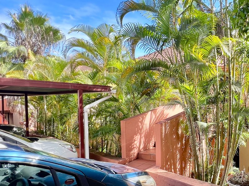 Exterior view of the apartment complex featuring a covered carport area and a tiled staircase leading to the entrance, surrounded by established palm trees.