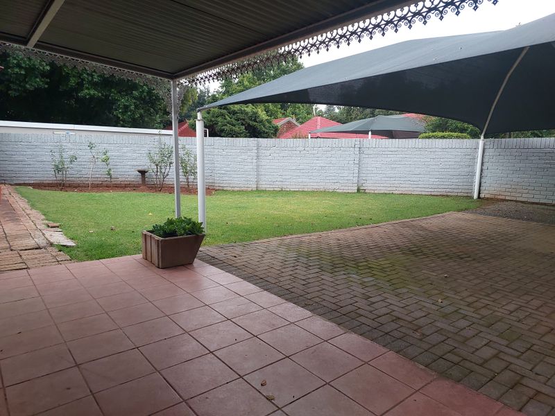 A covered patio area overlooking a private garden with a paved driveway, brick boundary wall, and shade sail for outdoor comfort.