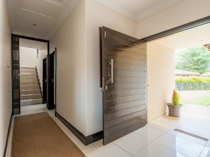 A bright and welcoming entrance hall featuring a modern dark wood pivot front door, tiled flooring, and a staircase leading to the upper level.