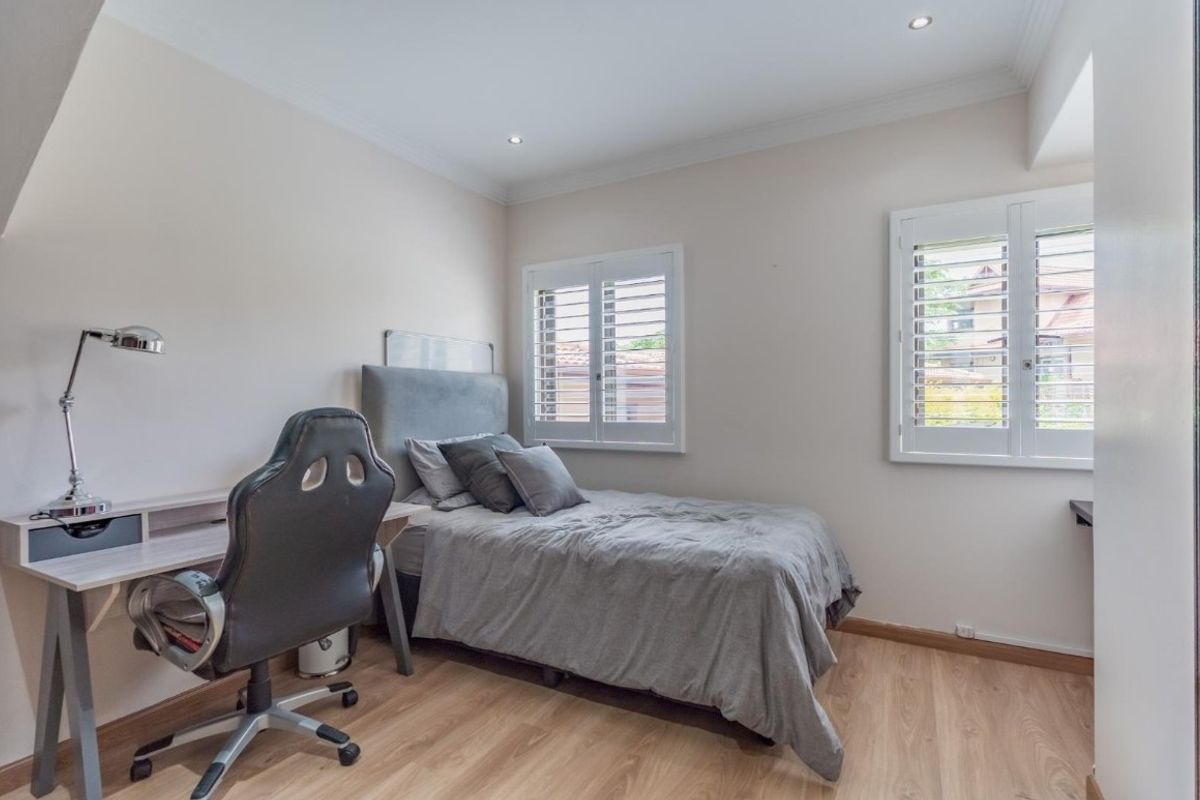 A well-maintained bedroom featuring light laminate flooring, recessed downlights, and plantation shutters, offering a clean and neutral living space.