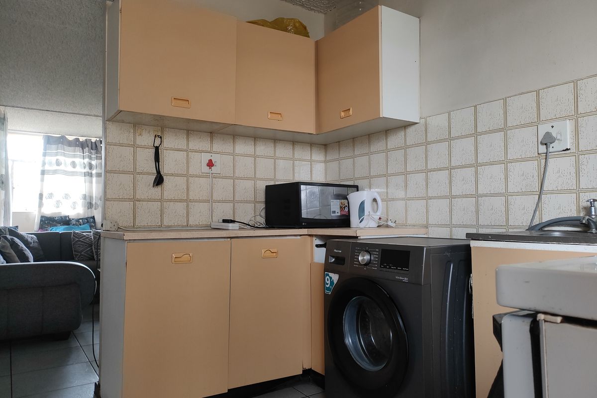 A compact kitchen area featuring dated cabinetry, tiled splashbacks, and integrated space for a washing machine, opening into the adjacent living area.