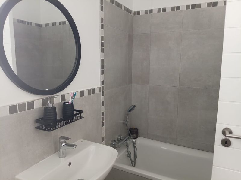 A modern bathroom featuring a white basin, a bathtub with a handheld shower mixer, and grey tiled walls with decorative mosaic accents.