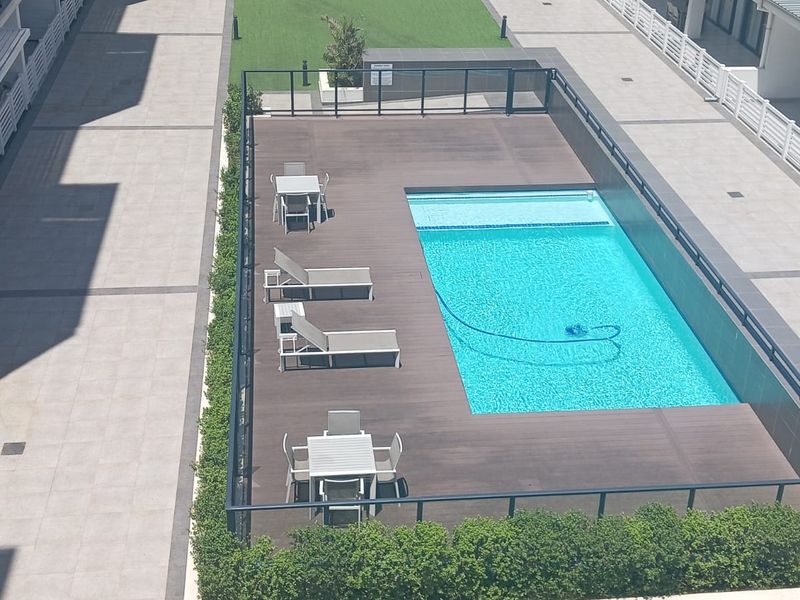 An elevated view of a modern apartment complex courtyard featuring a swimming pool with timber decking, paved walkways, and landscaped garden areas.