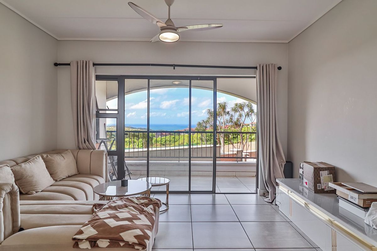 A bright living room featuring large sliding glass doors that open onto a balcony with scenic coastal views and a ceiling fan.