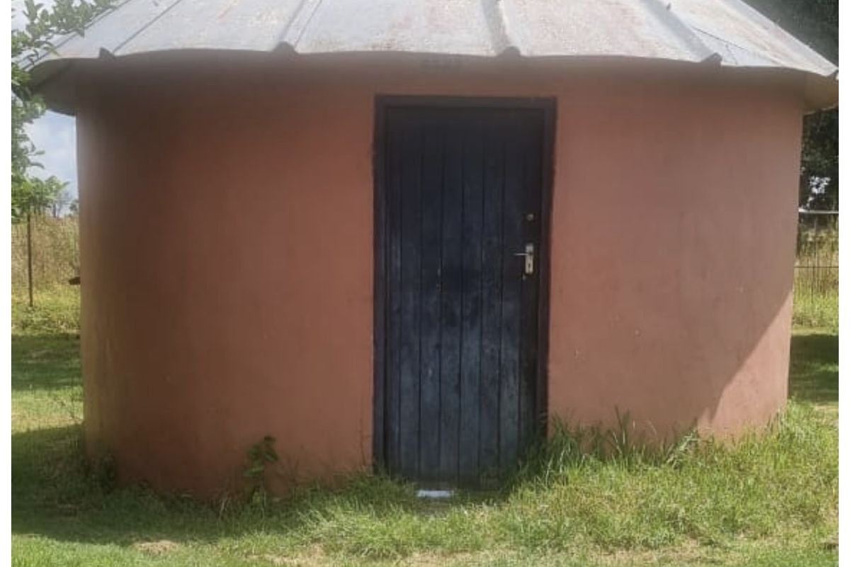 A small, circular outbuilding featuring a conical corrugated metal roof and a single timber entrance door, situated in a grassy farm garden setting.