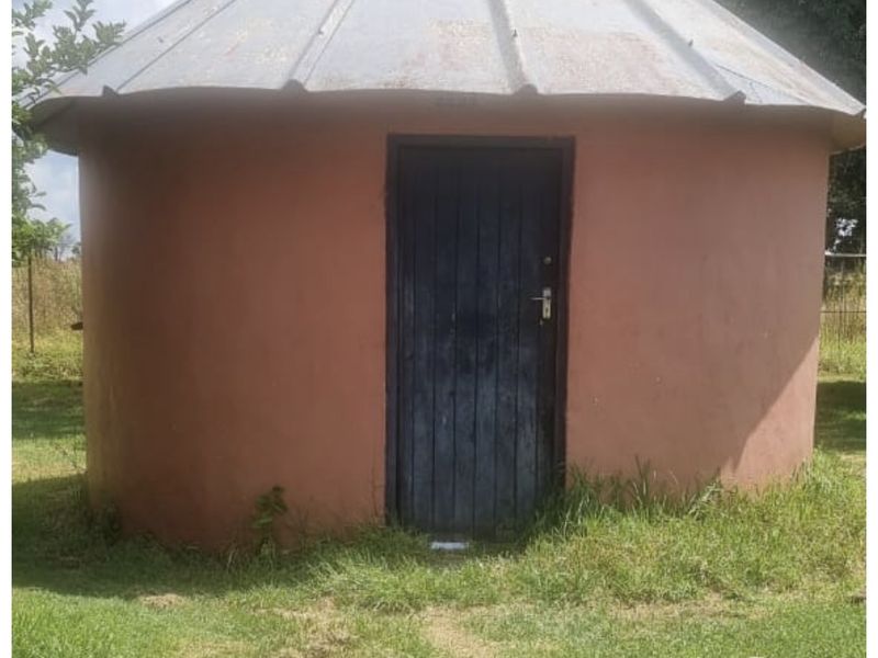 A small, circular outbuilding featuring a conical corrugated metal roof and a single timber entrance door, situated in a grassy farm garden setting.