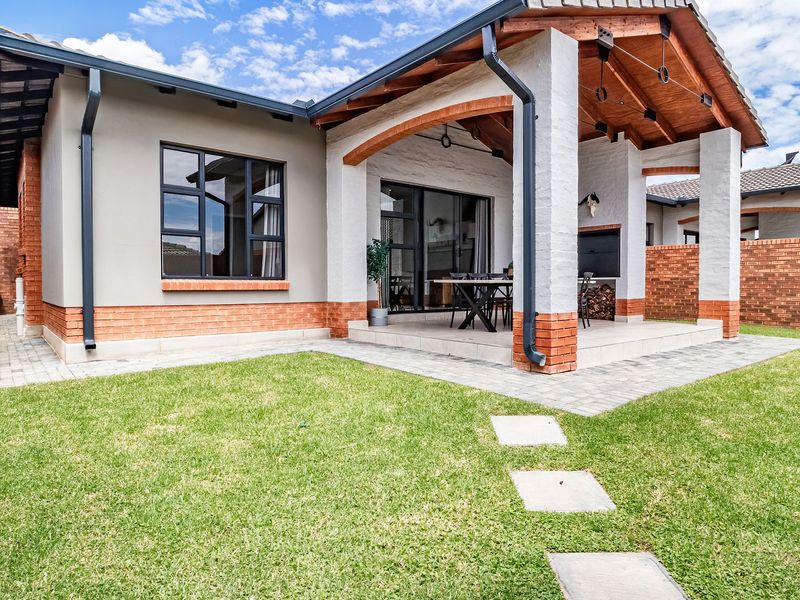 A well-maintained rear exterior view of a townhouse featuring a covered patio with a built-in braai, brick accents, and a landscaped garden.