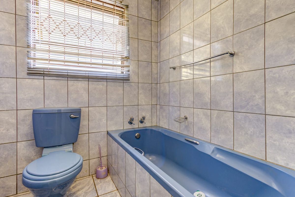 A dated bathroom featuring a blue bathtub and matching toilet, complemented by light blue wall tiles and a window with blinds.