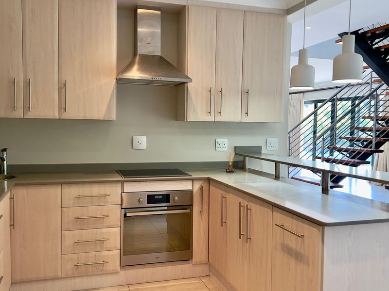 Modern kitchen featuring light wood cabinetry, stone countertops, integrated stainless steel appliances, and a breakfast bar overlooking an open-plan living area with staircase.