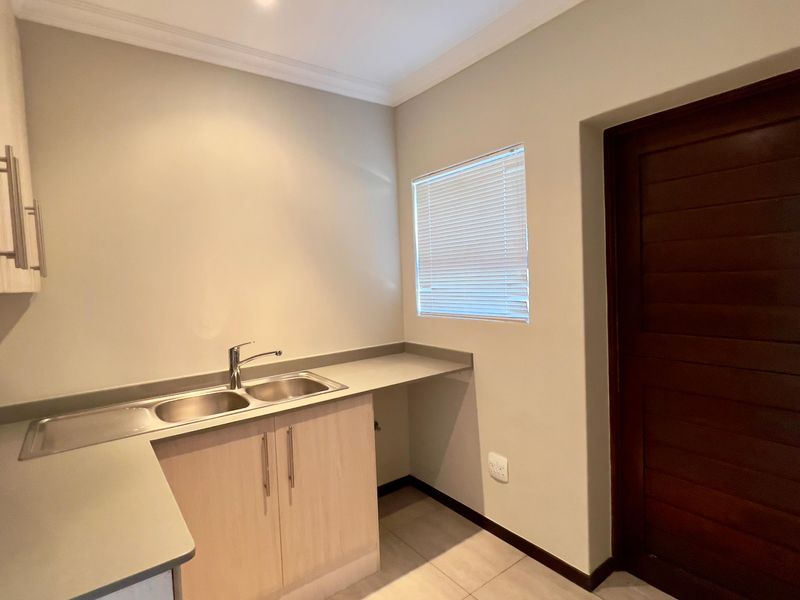 A clean and functional scullery featuring double bowl stainless steel sink, laminate cabinetry, and tiled flooring with direct access via a solid wood door.