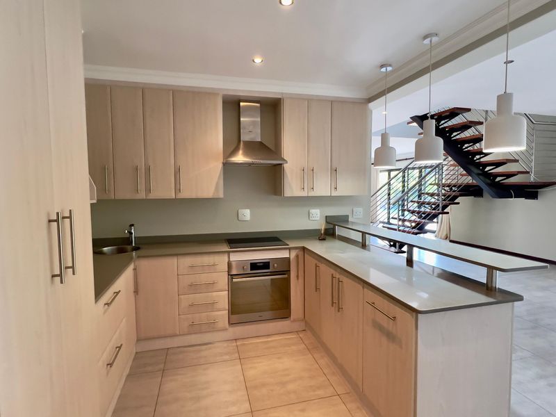 Modern kitchen featuring light wood cabinetry, stone countertops, integrated stainless steel appliances, and a breakfast bar overlooking the open-plan living area.