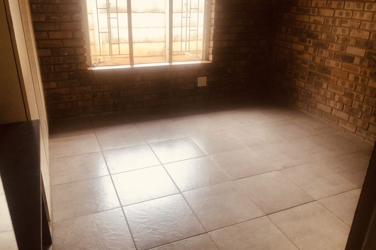 A vacant bedroom featuring exposed face-brick walls, tiled flooring, and a window fitted with security bars.