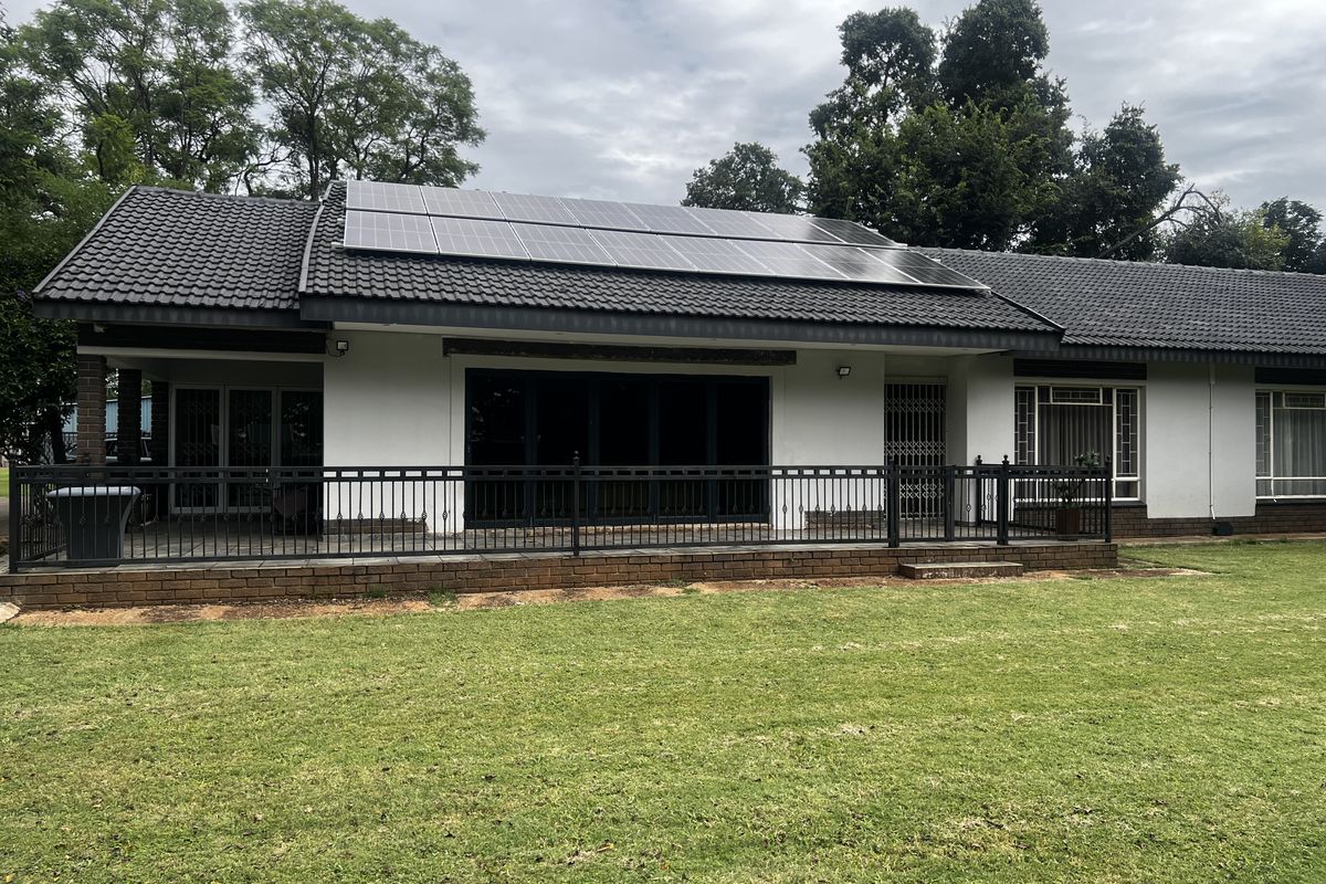 Rear view of the house featuring a covered patio, brick base, black metal railing, and roof-mounted solar panels overlooking a spacious lawn.