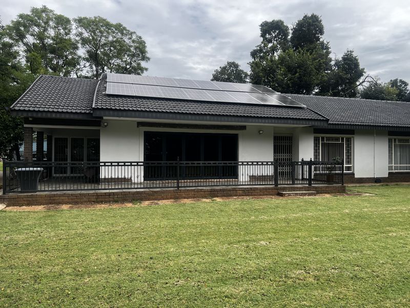 Rear view of the house featuring a covered patio, brick base, black metal railing, and roof-mounted solar panels overlooking a spacious lawn.