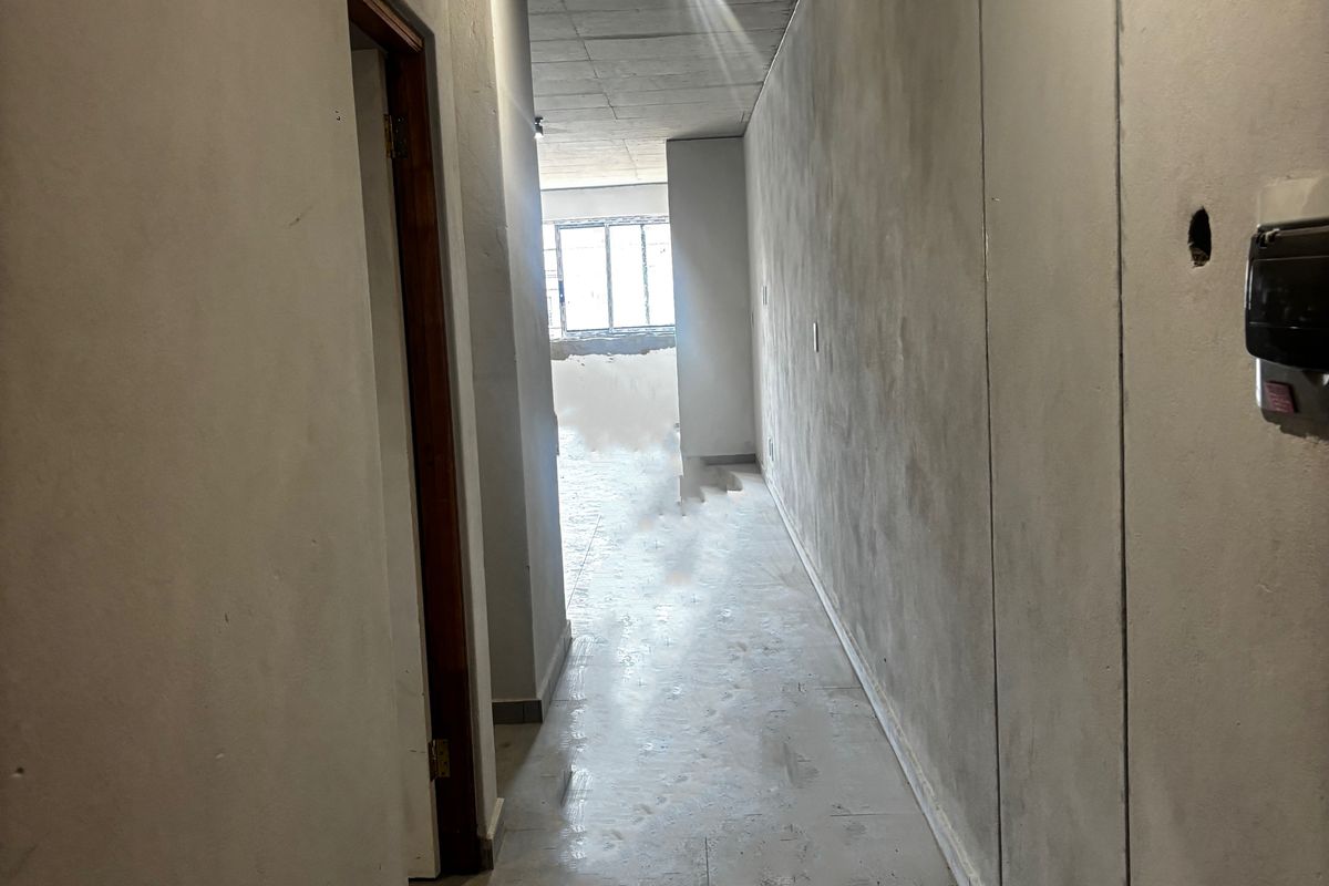 A hallway in an unfinished apartment, featuring raw concrete ceilings, unpainted plastered walls, and tiled flooring, awaiting final interior finishes.