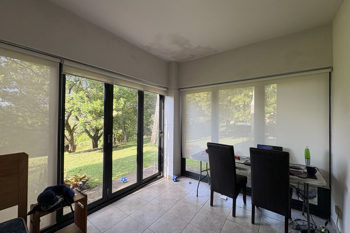 A bright living area featuring large floor-to-ceiling glass sliding doors with roller blinds, tiled flooring, and views of the surrounding garden.