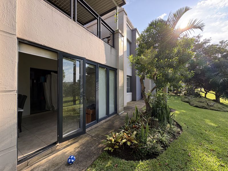 Rear view of the apartment featuring a paved patio area, landscaped garden, and large glass sliding doors providing seamless indoor-outdoor flow.