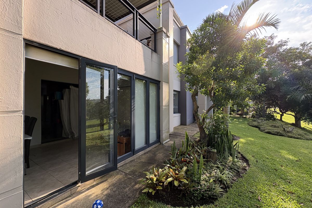 Rear view of the apartment featuring a paved patio area, landscaped garden, and large glass sliding doors providing seamless indoor-outdoor flow.