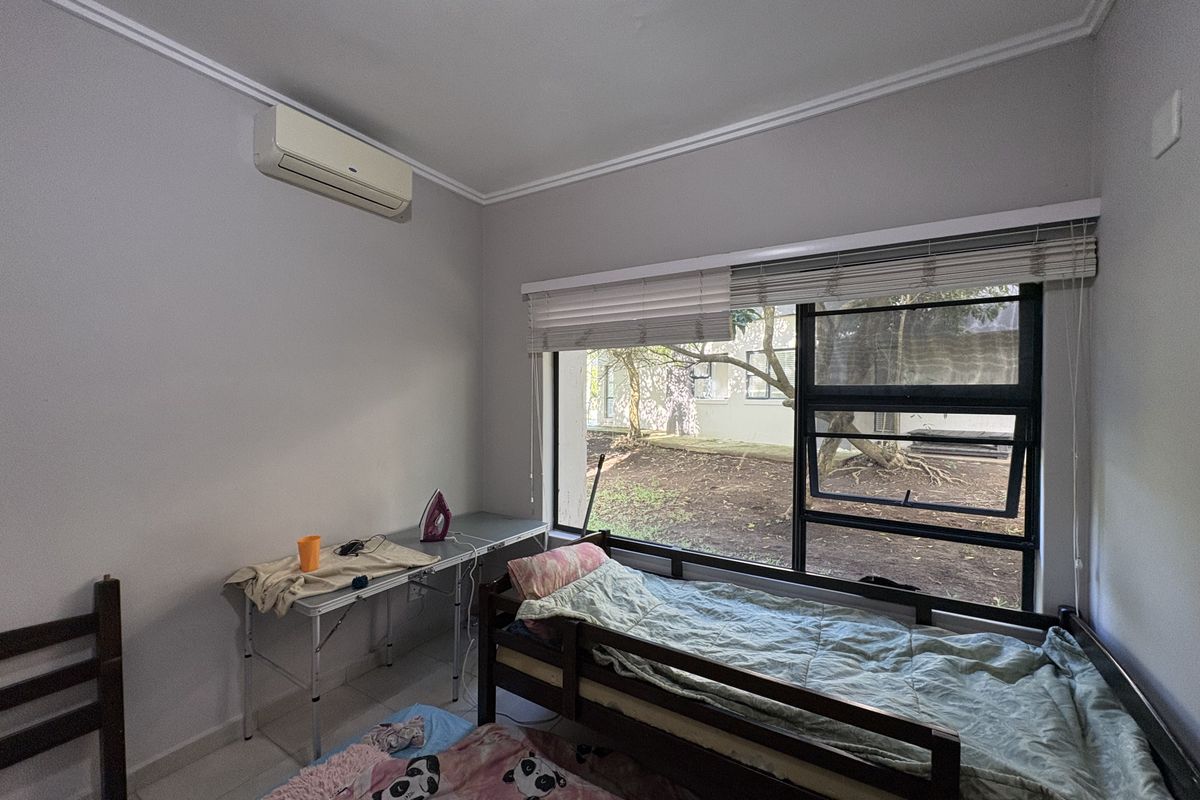 A bedroom featuring a large window with blinds, tiled flooring, and a wall-mounted air conditioning unit, offering a view of the exterior grounds.