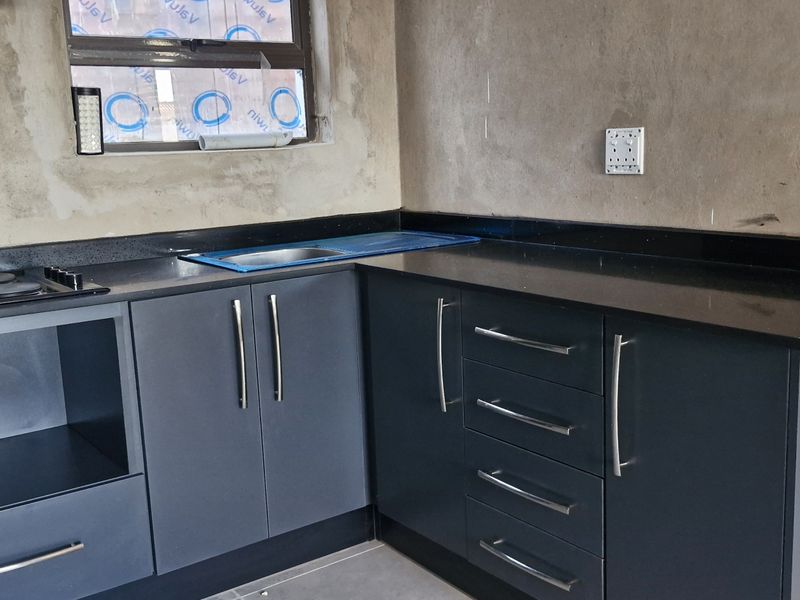 A partially completed kitchen featuring dark grey cabinetry, black granite countertops, and an integrated electric hob, currently awaiting final wall finishes.
