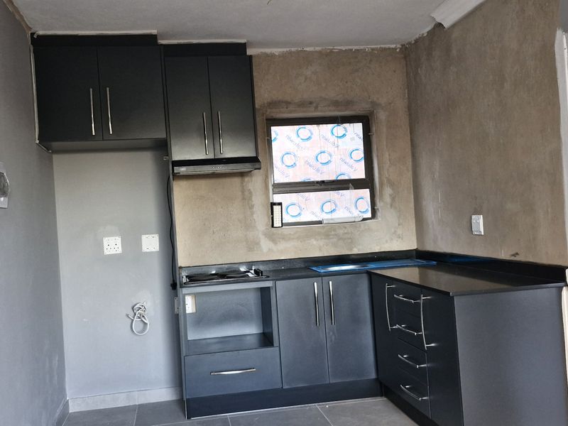 A compact kitchen featuring dark cabinetry, black stone countertops, and a built-in hob, currently requiring final wall finishes and minor electrical completion.