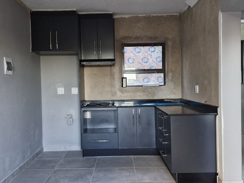 A compact kitchen featuring dark built-in cabinetry, stone countertops, and a hob, currently requiring final wall finishing and plastering.