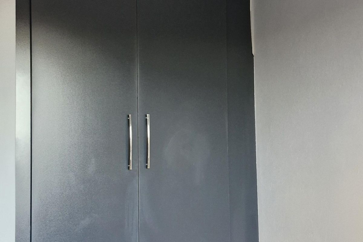 A clean, empty bedroom space featuring a modern dark grey built-in cupboard, neutral tiled flooring, and decorative ceiling cornicing.