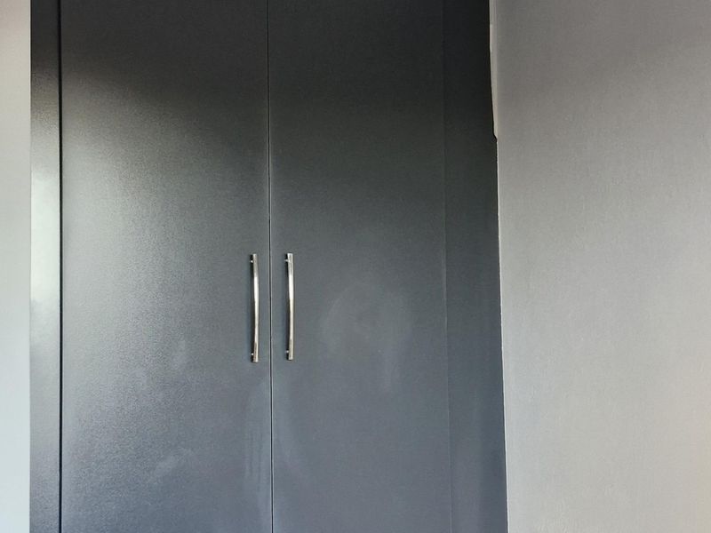 A clean, empty bedroom space featuring a modern dark grey built-in cupboard, neutral tiled flooring, and decorative ceiling cornicing.