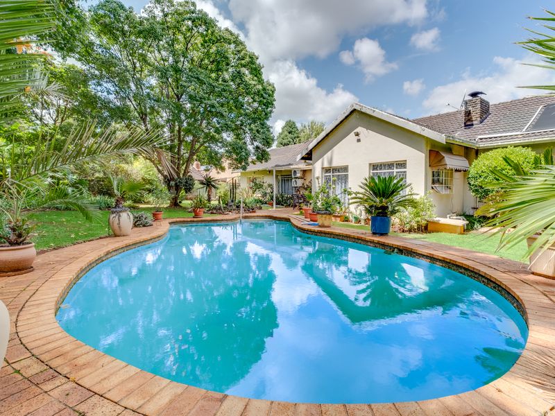 A well-maintained rear garden featuring a swimming pool with brick coping, surrounded by established trees and landscaped greenery.