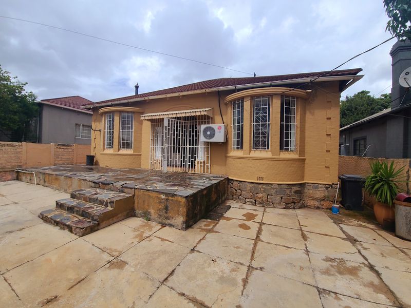 Rear view of the house featuring a stone-clad patio, bay windows, and an exterior air conditioning unit, set against a paved backyard area.