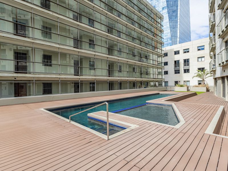 A communal outdoor pool area featuring a modern swimming pool surrounded by composite decking and overlooked by the apartment building's glass facade.