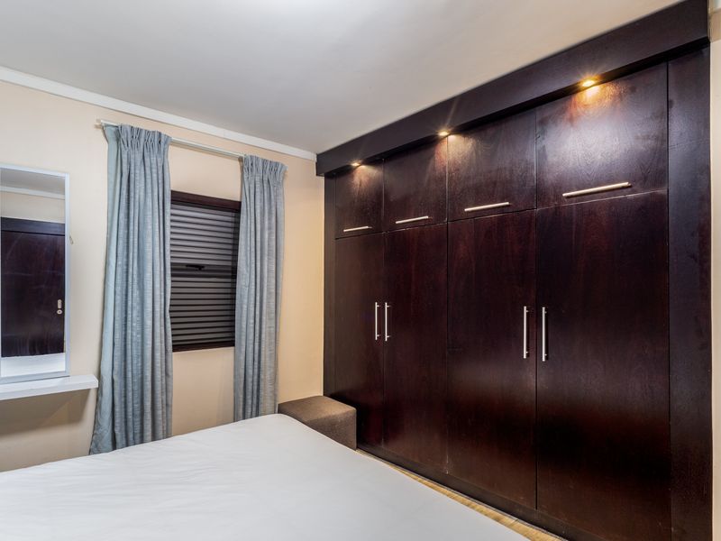 A well-maintained bedroom featuring dark wood built-in cupboards with integrated downlighting and a window fitted with a security roller shutter.