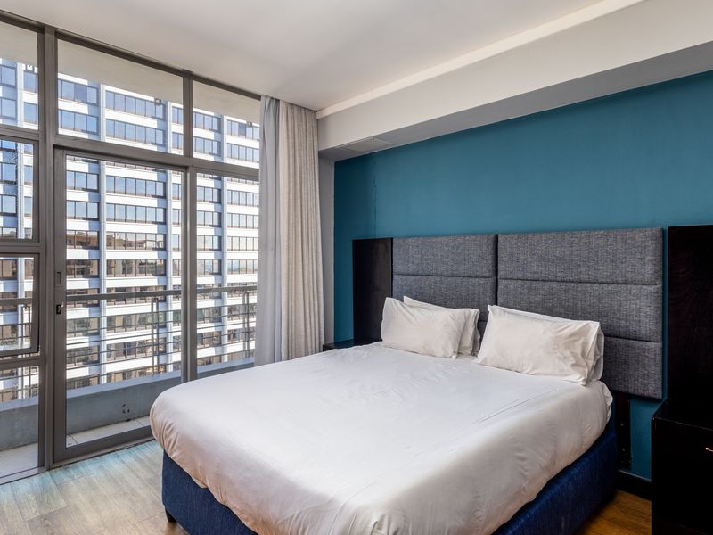 Modern bedroom featuring floor-to-ceiling windows with balcony access, a built-in headboard, and laminate flooring, offering an urban city view.