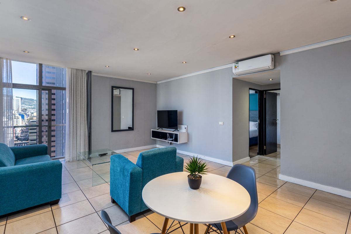 A bright open-plan living area featuring tiled floors, recessed lighting, a wall-mounted air conditioning unit, and access to a balcony with city views.