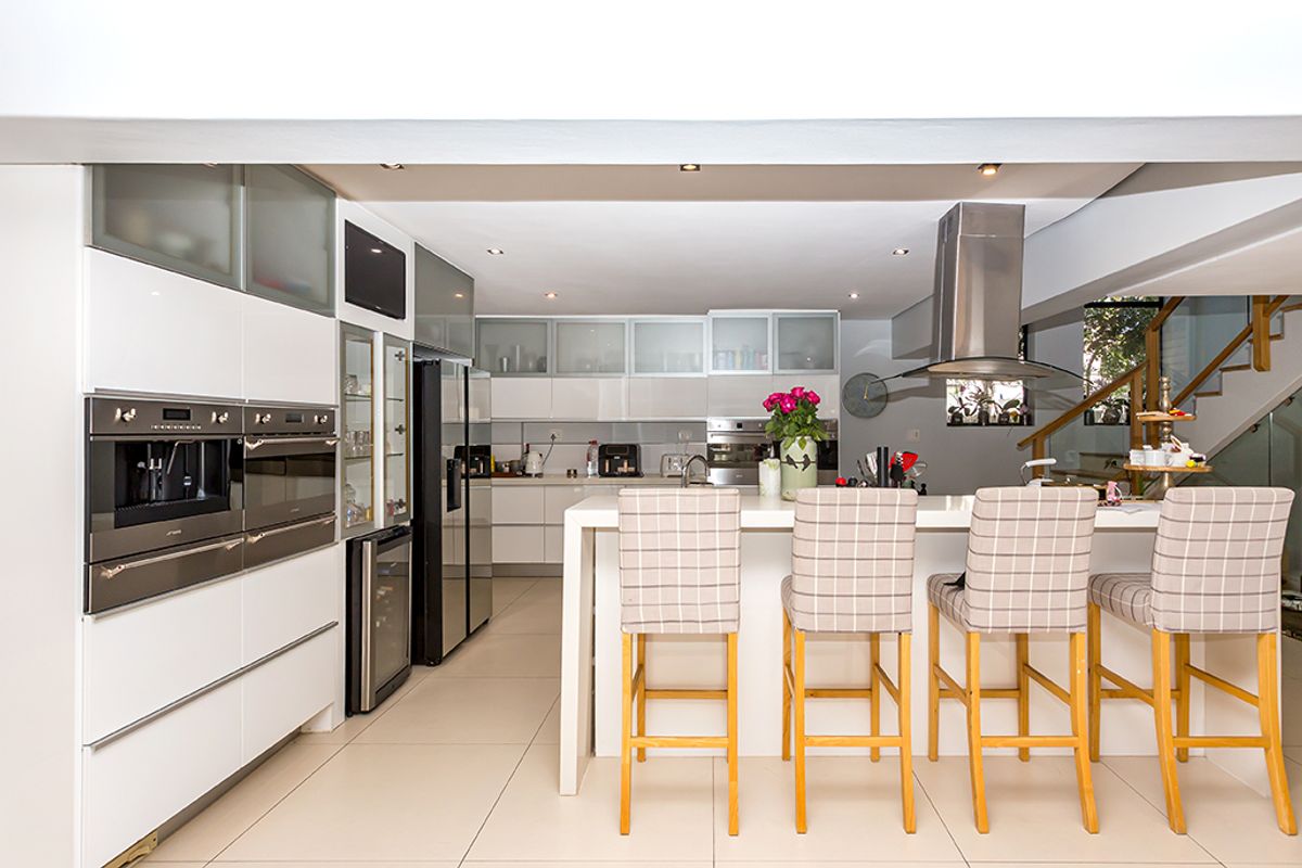 Modern open-plan kitchen featuring sleek white cabinetry, integrated stainless steel appliances, a central island with breakfast bar seating, and recessed ceiling lighting.