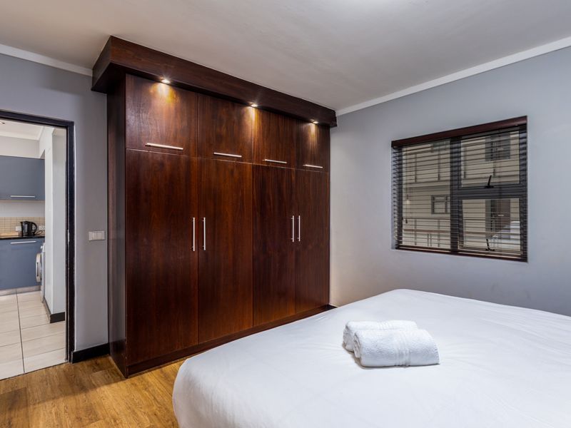 A well-maintained bedroom featuring dark wood built-in wardrobes with integrated lighting, laminate flooring, and a view into the adjacent kitchen area.