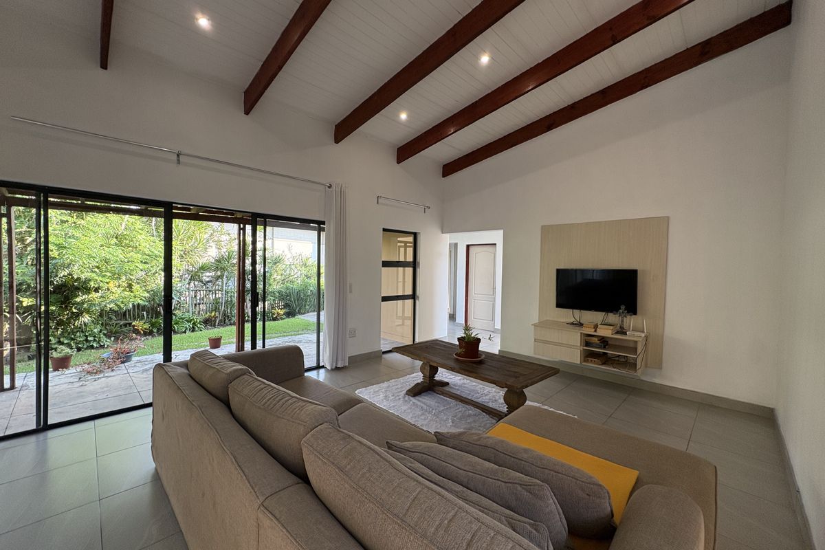 Spacious living room featuring high vaulted ceilings with exposed timber beams, large sliding glass doors opening to a garden patio, and tiled flooring.