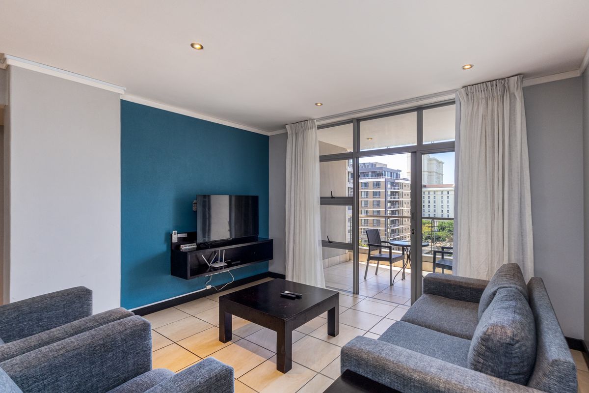 A well-lit living room featuring tiled floors, a bold blue accent wall, and direct access to a private balcony through large sliding glass doors.